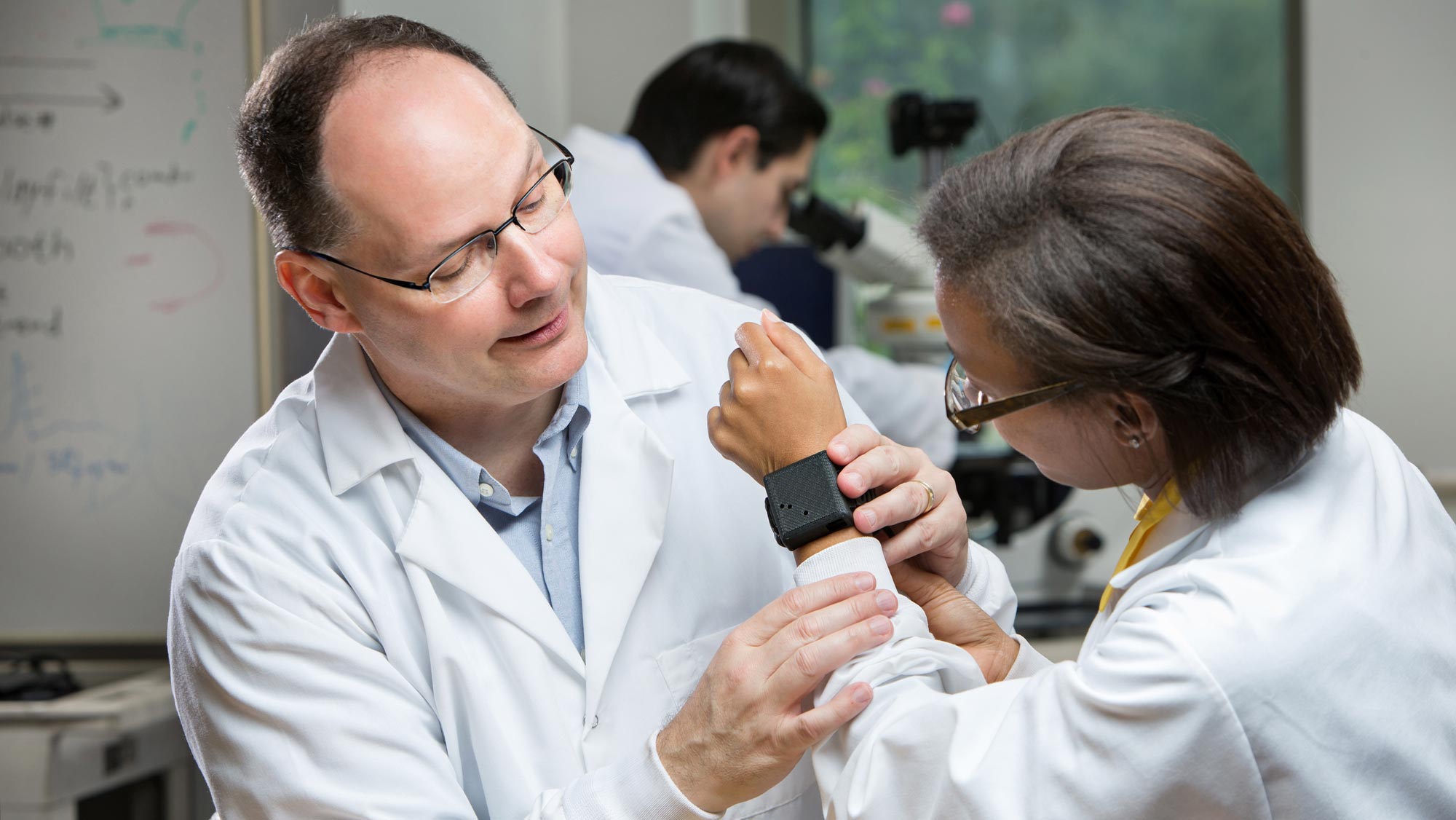 A man with glasses in a white lab coat affixes a wrist device that resembles a smartwatch to a young woman with glasses in a white lab coat and with her back to the camera.